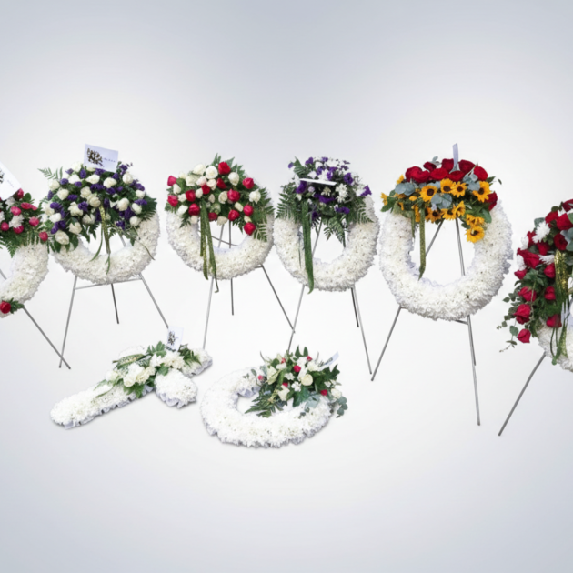 Large collection of assorted funeral flower arrangements including wreaths, a heart, and a cross in white and purple.